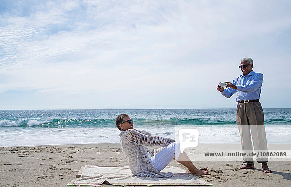 Senior couple on beach  man taking photograph of woman using smartphone