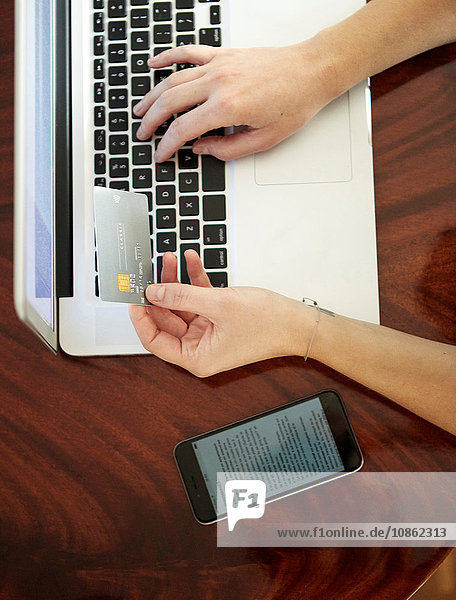 Draufsicht einer Frau mit Smartphone  die mit einem Laptop eine Kreditkarte hält