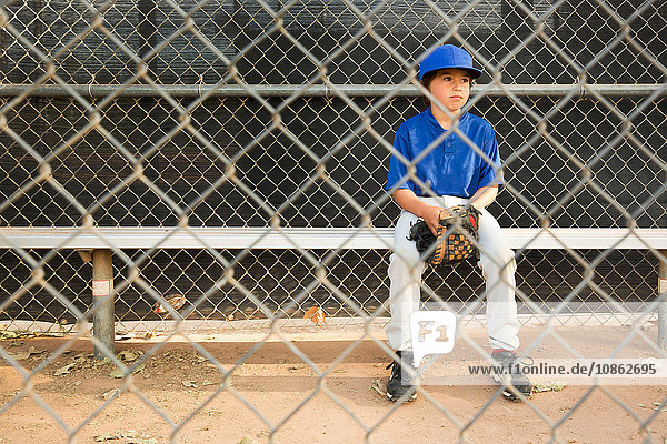 Boy sitting on bench behind wire fence at baseball practise