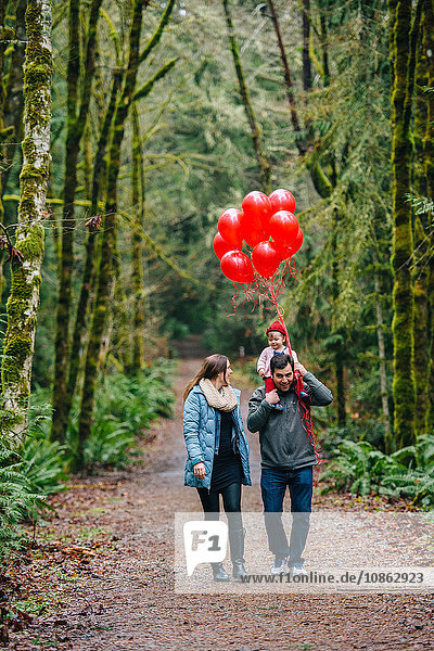 Mittelgroßes erwachsenes Paar trägt Kleinkind-Tochter auf den Schultern mit einem Haufen roter Luftballons im Wald