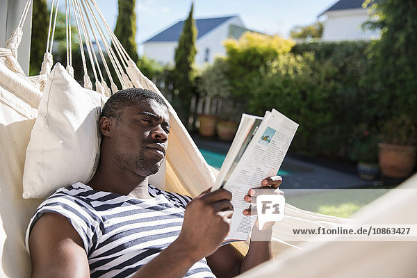 Auf Gartenhängematte liegender junger Mann liest eine Zeitschrift