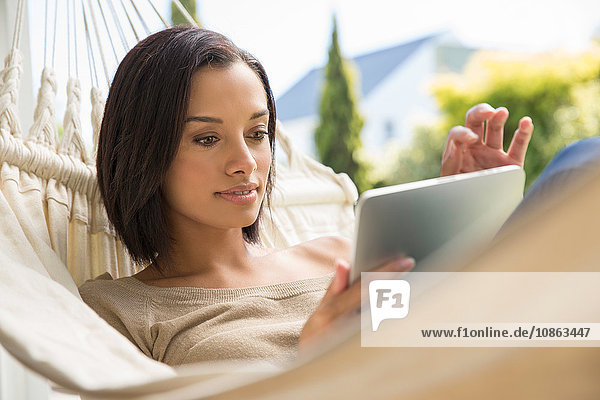 Junge Frau liegt auf Gartenhängematte und benutzt Touchscreen auf digitalem Tablett