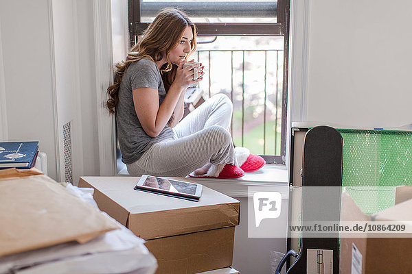 Junge Frau sitzt auf dem Fensterbrett eines neuen Hauses  hält heißes Getränk  Kartons im Zimmer