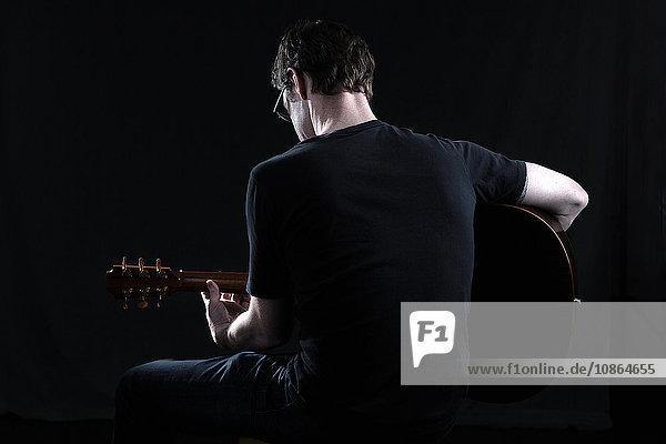 Low key rear view of mid adult man playing acoustic guitar