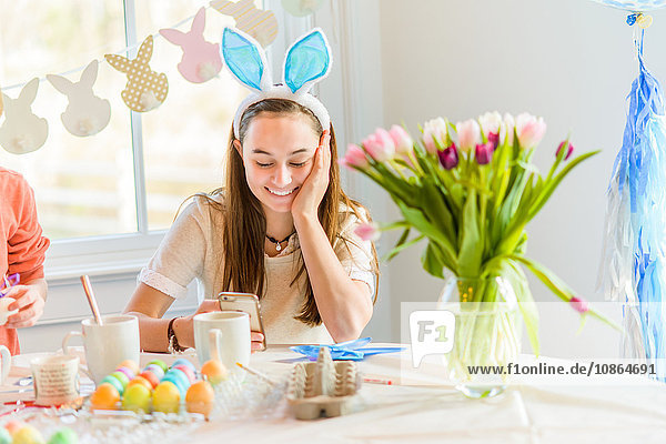 Teenager-Mädchen liest am Tisch Smartphone-Texte und färbt hart gekochte Eier für Ostern