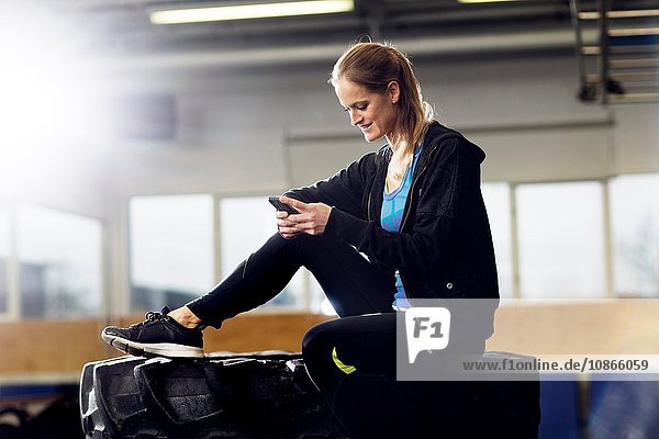 Weibliche Crossfitterin sitzt auf einem Reifenstapel im Fitnessstudio und liest Smartphone-Texte