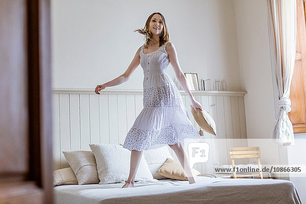 Junge Frau in langem weißen Kleid springt auf Bett und schaut lächelnd in die Kamera
