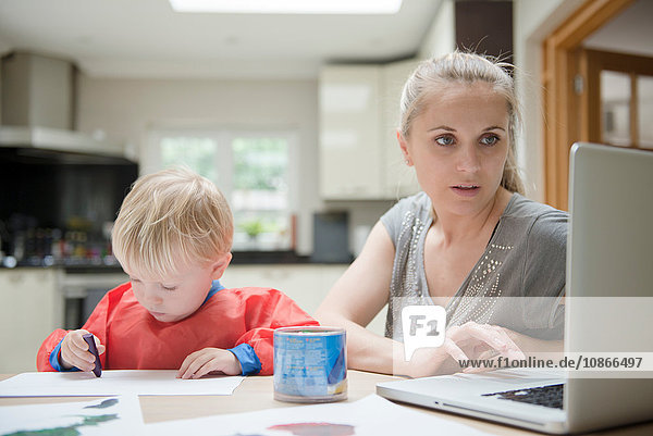 Mutter benutzt Laptop  während der Sohn neben ihr auf Papier zeichnet