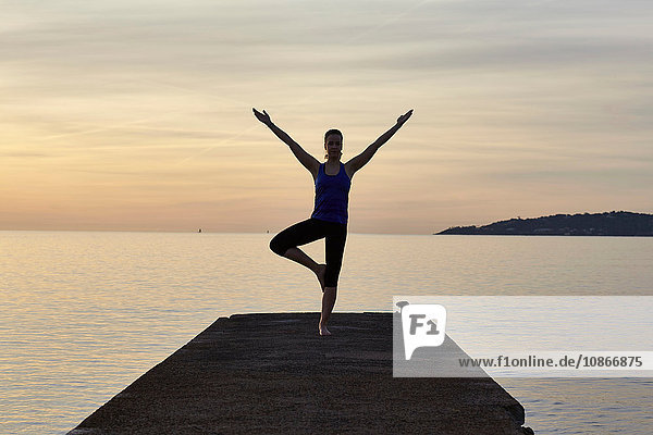 Junge Frau am Pier stehend  in Yogastellung  bei Sonnenuntergang