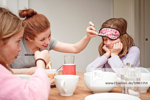 Mittlere erwachsene Frau am Frühstückstisch  die unter die Schlafmaske ihrer Töchter schaut