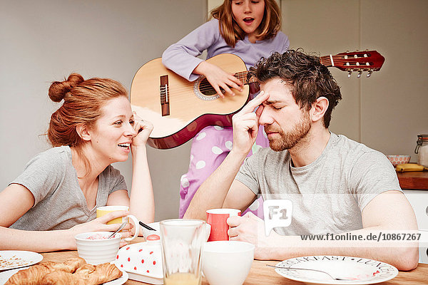Müde mittlere erwachsene Eltern am Frühstückstisch  während die Tochter Gitarre spielt