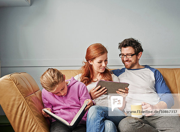 Mid adult parents and daughter on sofa on reading book and digital tablet