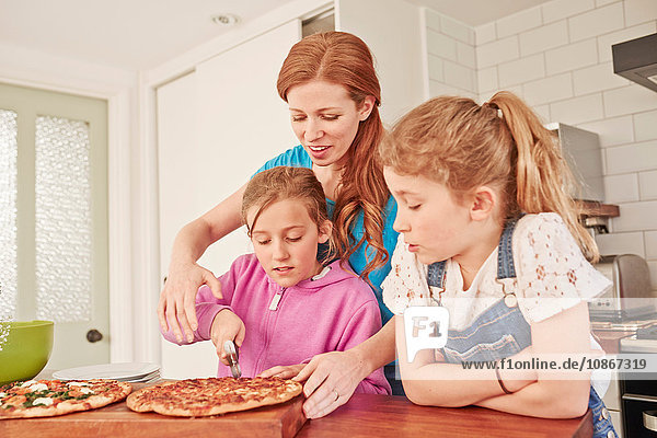 Mittlere erwachsene Frau schneidet Pizza für Töchter an der Küchenbank