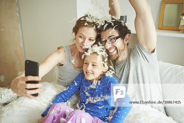 Mittelgroßes erwachsenes Paar und Tochter mit Kissenschlachtenfedern bedeckt  die Smartphone-Selfie im Bett nehmen