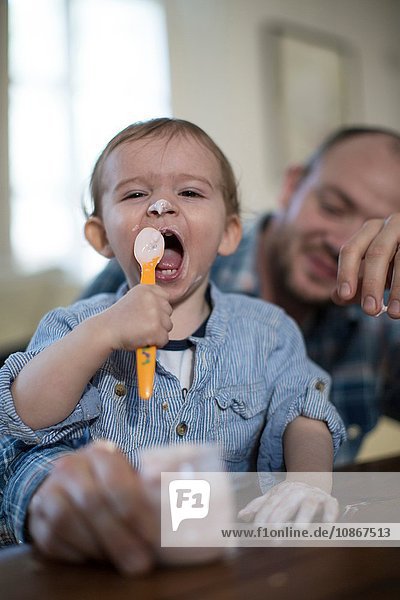 Kleiner Junge füttert sich selbst mit einem Löffel Joghurt und schaut in die Kamera  den Mund geöffnet