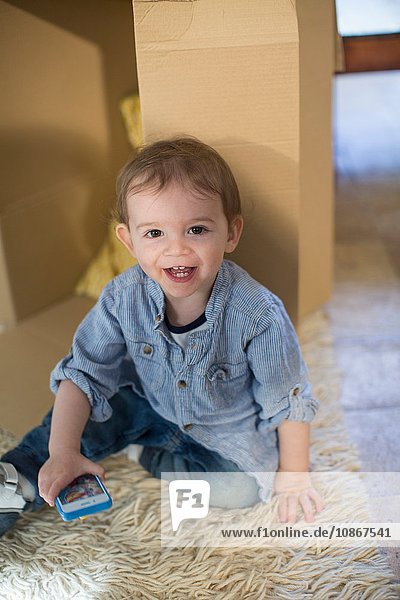 Kleiner Junge sitzt vor Pappkarton mit Smartphone und schaut lächelnd in die Kamera