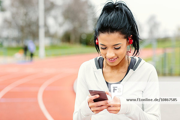 Young woman beside running track  holding smartphone  wearing earphones  smiling