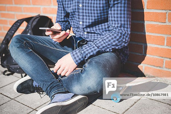 Neck down view of young male urban skateboarder sitting on sidewalk reading smartphone text