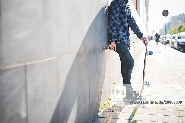 Junger männlicher Skateboarder  der sich an die Gehsteigwand lehnt