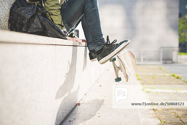 Taille nach unten Ansicht eines jungen männlichen Skateboardfahrers  der an der Wand sitzt und das Skateboard mit den Füßen dreht.