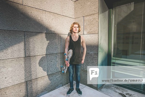 Portrait of young male urban skateboarder standing in corner holding skateboard