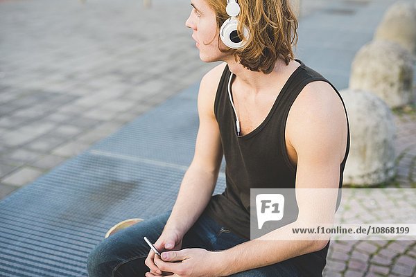 Junger Mann sitzt auf dem Bürgersteig und hört Kopfhörermusik.