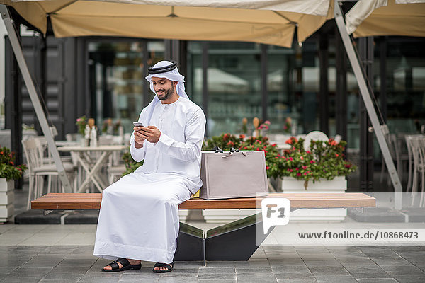 Männlicher Einkäufer in traditioneller Kleidung aus dem Nahen Osten sitzt auf Bank und liest Smartphone-Text  Dubai  Vereinigte Arabische Emirate