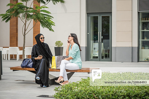 Junge Frau aus dem Nahen Osten in traditioneller Kleidung mit Freundin auf Bank sitzend  Dubai  Vereinigte Arabische Emirate
