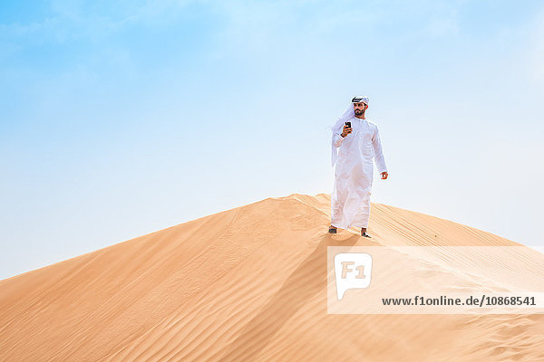 Mann aus dem Nahen Osten trägt traditionelle Kleidung mit Smartphone auf Wüstendüne  Dubai  Vereinigte Arabische Emirate