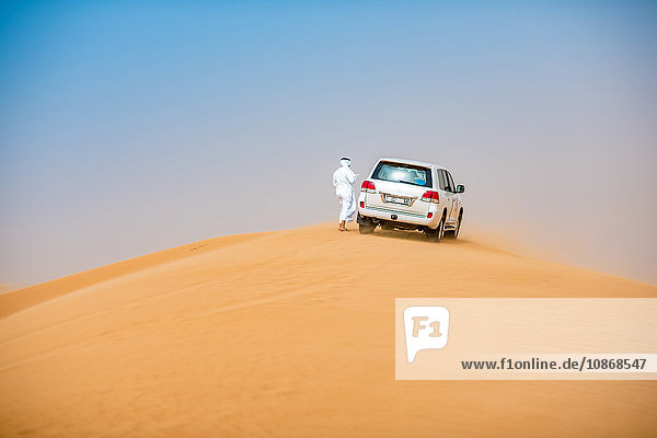 Mann aus dem Nahen Osten in traditioneller Kleidung mit Geländewagen auf Wüstendüne geparkt  Dubai  Vereinigte Arabische Emirate
