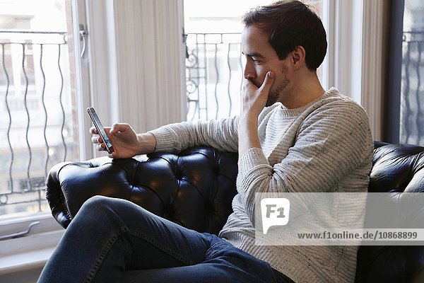 Mittelgroßer erwachsener Mann sitzt auf dem Sofa und schaut auf ein Smartphone