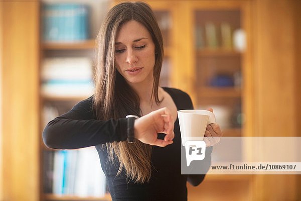 Young woman at home  holding hot drink  looking at watch