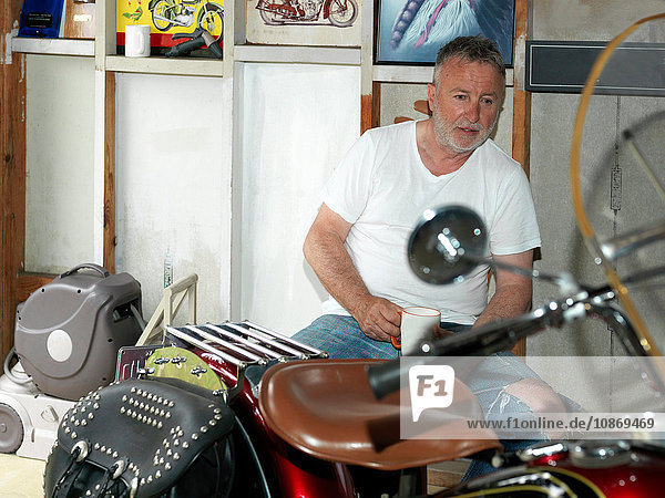 Älterer Mann sitzt mit einem Kaffee und starrt auf ein Motorrad in der Garage