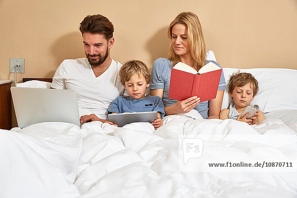 Mutter und Vater im Bett mit Söhnen  die mit Technik und Lesebuch umgehen
