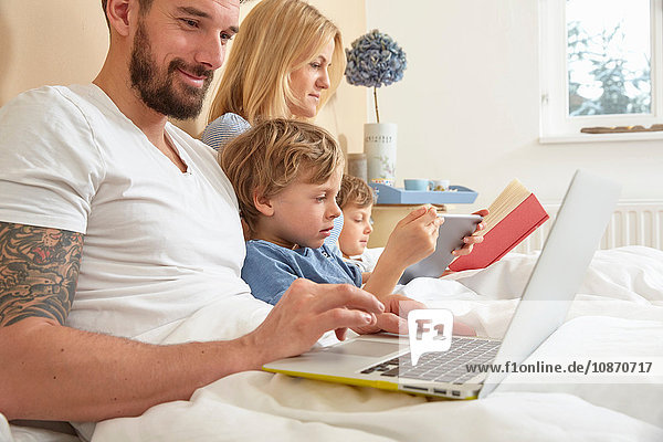 Mother and father in bed with sons using technology  reading book