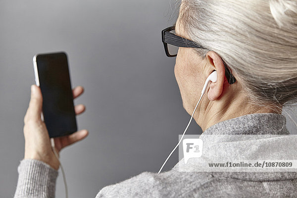Über-Schulter-Ansicht einer grauhaarigen Frau  die Ohrstöpsel trägt und ein Smartphone hält