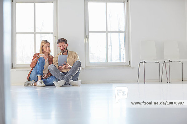 Paar sitzt auf dem Boden und stöbert auf einem digitalen Tablet in einem leeren neuen Haus