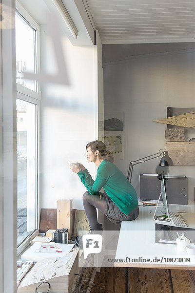 Blick durch Glas  Seitenansicht in voller Länge einer reifen Frau  die auf einem Schreibtisch sitzt und aus dem Fenster schaut
