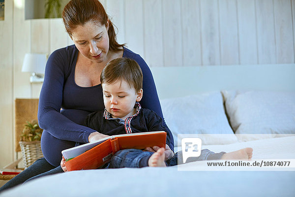 Schwangere Mutter und kleiner Junge sitzen zusammen auf dem Bett und lesen Buch