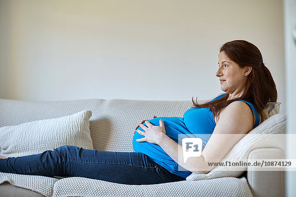 Seitenansicht einer schwangeren Frau  die den Bauch hält  sich auf dem Sofa entspannt und lächelnd wegschaut