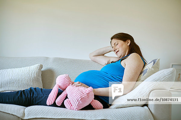 Seitenansicht einer schwangeren Frau mit rosa Plüschtier in der Hand  die Augen geschlossen und auf dem Sofa ruhend