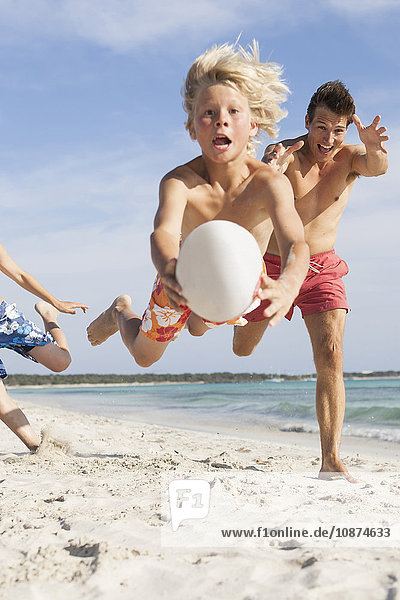 Junge springt in der Luft mit einem Rugbyball  der von Bruder und Vater am Strand verfolgt wird  Mallorca  Spanien