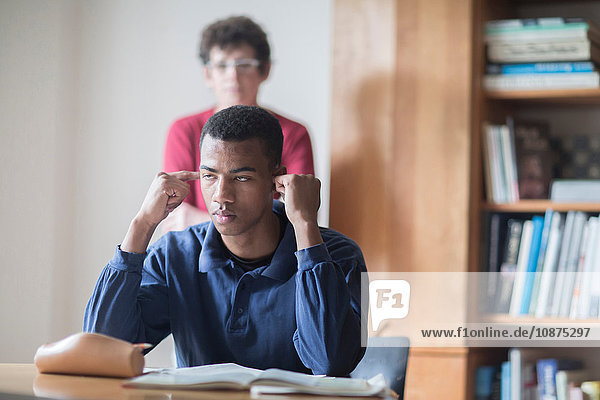 Junger männlicher Gymnasiast am Schreibtisch sitzend mit Fingern in den Ohren und Lehrer dahinter stehend