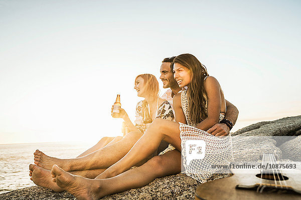Drei mittelgroße erwachsene Freunde sitzen bei Sonnenuntergang mit Flaschenbier am Strand Kapstadt Südafrika