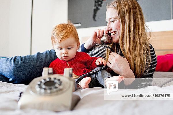Mittelgroße erwachsene Frau und kleine Tochter im Bett sprechen am Festnetztelefon