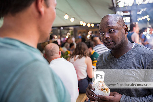 Male customers chatting and eating burgers at cooperative food market stall