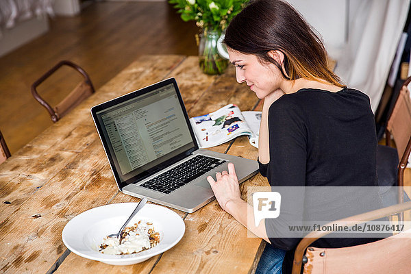 Junge Frau sitzt am Tisch in Wohnung mit Müslifrühstück  liest Laptop