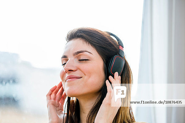Beautiful young woman listening to headphone music in front of apartment window