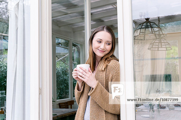 Woman leaning against doorway holding coffee cup looking away smiling
