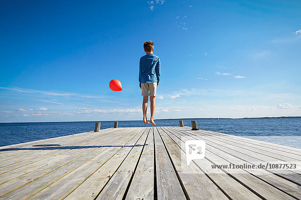 Junge springt auf Holzsteg  hält roten Heliumballon in der Hand  Rückansicht